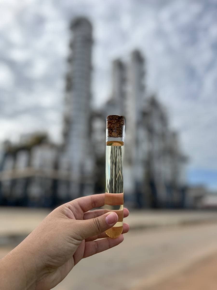Mão segurando tubo de ensaio com amostra, com a unidade industrial desfocada ao fundo